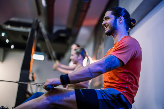 Side profile view of a muscular man focused on his form while exercising on a rowing ergometer next to other people in a dark, atmospheric gym.