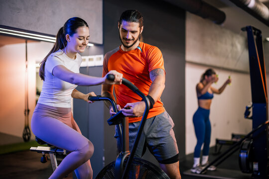 A male personal trainer stands over a female client, giving instructions and correcting her form on an indoor rowing machine.
