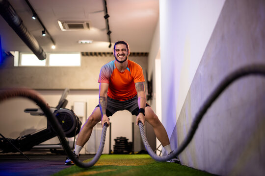 Wide angle view of a determined male athlete in an orange t-shirt slamming heavy ropes, focusing on core stability and upper body strength.