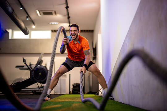 Full body shot of a strong man in a red t-shirt executing perfectly symmetrical waves with heavy battle ropes during a high-intensity functional training session.