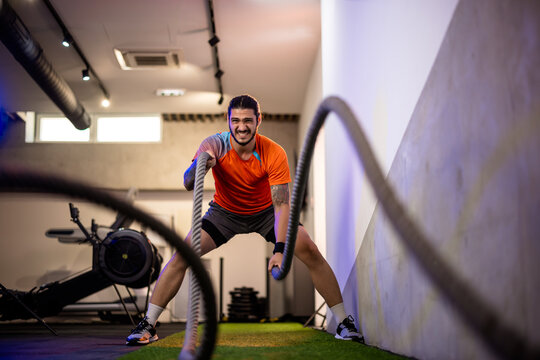 Front view of a focused male fitness trainer slamming heavy battle ropes on the floor, creating a dynamic wave effect in a modern functional training zone.