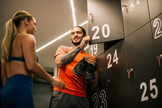 A young man and a woman in sportswear sharing a fun, casual conversation by the lockers, holding sports equipment and enjoying their time after a workout.