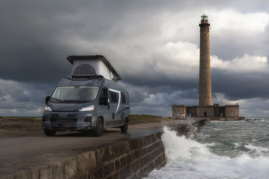 View of a modern camper van parked on a stone causeway near the historic Gatteville-le-Phare lighthouse with crashing waves under a dramatic cloudy sky in Gatteville-le-Phare, Normandy, France.