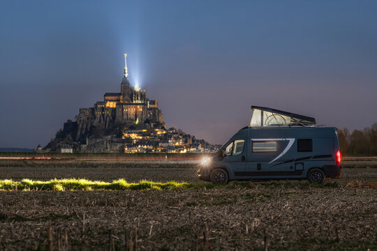 View of a camper van parked in a field at night with the illuminated Mont Saint-Michel abbey in the background in Mont Saint-Michel, Normandy, France.