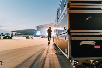Naklejka premium Black touring road cases lined up backstage with a lone crew member walking toward a luminous blue stage for live event production services