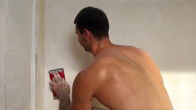 Young man uses brush to clean finished wall surface removing dust and small imperfections after renovation work. Male worker carefully prepares the smooth wall for next finishing stage inside room