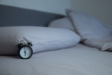 On the light gray bed linen there is a black miniature alarm clock against the background of two soft pillows in the dim lighting. Concept: the feeling of early morning, calm bedroom interior. High © Ксения Мнасина