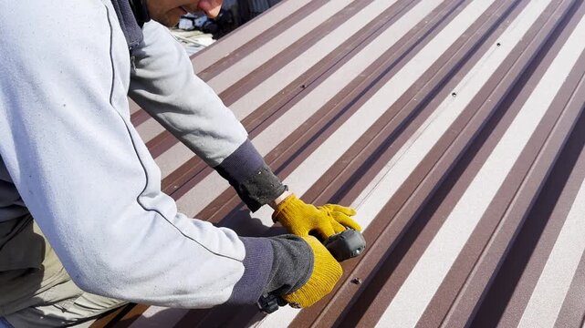 Male hands in gloves use cordless screwdriver to fasten roofing sheets in house roof. Young worker secures screws into surface performing careful installation work during residential construction