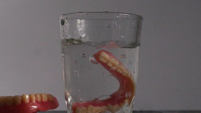 A removable full denture falls into a glass of water causing a splash, while another one lies on the table. Demonstration of daily hygienic care and cleaning process for false teeth at home