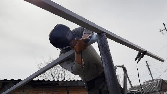 Manual laborer in protective mask weld iron detail on carport frame. Professional young repairman with welding machine working at construction. Male welder operating with metal at the site. Close up