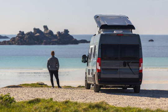 View of a man standing next to a grey camper van with a pop-up roof parked on a gravel path overlooking a sandy beach and rocky coastline in Normandy, France.