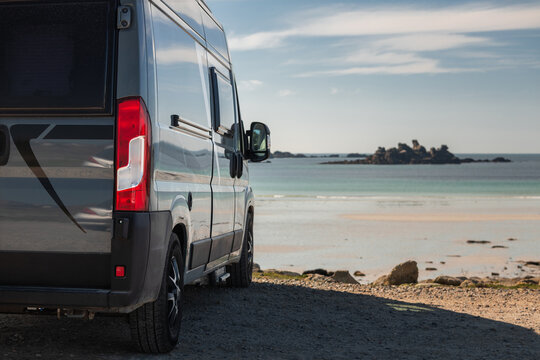 View of a grey camper van parked on a coastal road overlooking a sandy beach with rocky islets in the distance in Normandy, France.