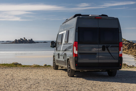 View of a grey camper van parked on a gravel path overlooking the sea with rocky outcrops in the distance in Normandy, France.