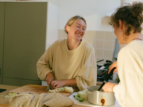 Young adult woman with a hearing aid chopping vegetables in a lived-in kitchen, smiling during a natural moment for healthy lifestyle and plant based content