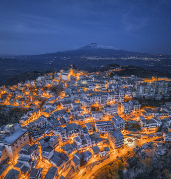 Aerial view of the historic town of Centuripe illuminated at night with Mount Etna in the background, featuring dense architecture and warm street lights in Centuripe, Sicily, Italy.