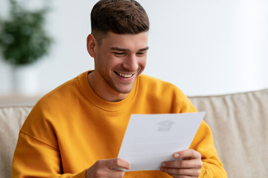 Smiling young man reads a letter while sitting on a sofa at home. Good news, personal success, positive result.