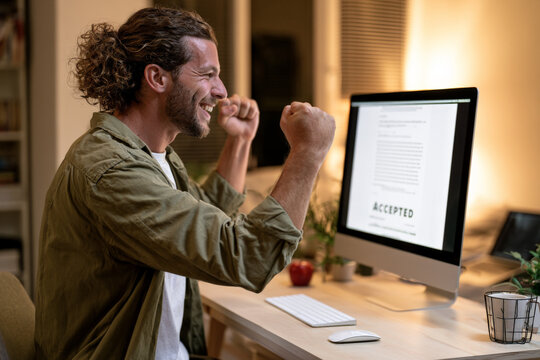 Bearded man celebrates beside a computer displaying an accepted document. Online approval, career success, positive result.