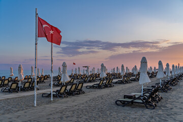 Fototapeta premium Beach chairs and umbrellas beneath a Turkish flag during a Mediterranean sunset in Antalya, Turkey.