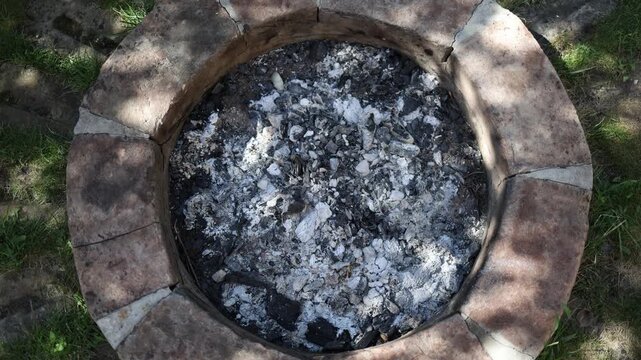 Cold gray ash and burned charcoal remain inside a round brick fire pit after an outdoor fire showing the quiet aftermath of backyard cooking camping and summer evenings