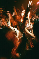 Fototapeta premium Crowded nightclub dancers raise their hands under warm orange lights, captured with energetic motion blur from a close perspective. Motion blur, selective focus.