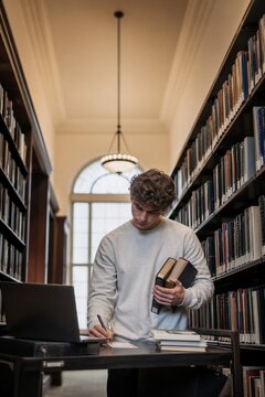 Student man verifying information with books and laptop in library. Researcher searching for data in archive.