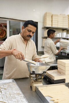Confectioner weighing indian sweets on scale in kitchen. Man working in bakery shop packing traditional mithai into paper box for customer. Small business food production process.