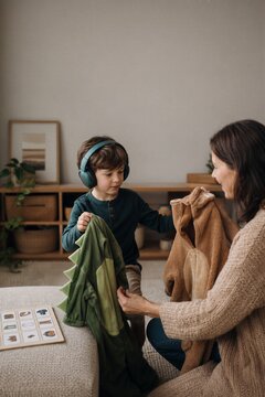 Woman mother helping young boy child choose between animal costumes for Halloween. Kid wearing headphones holding dinosaur outfit at home.