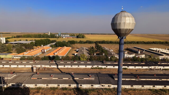 Aerial view of a farm featuring a prominent spherical water tower, long agricultural buildings with orange and grey roofs, and vast surrounding fields in Dolovo, Vojvodina, Serbia.