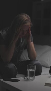 A woman in her thirties sits on a bed in a dark room, holding insomnia pills and a glass of water, looking troubled as sleep remains elusive and the night stretches on.