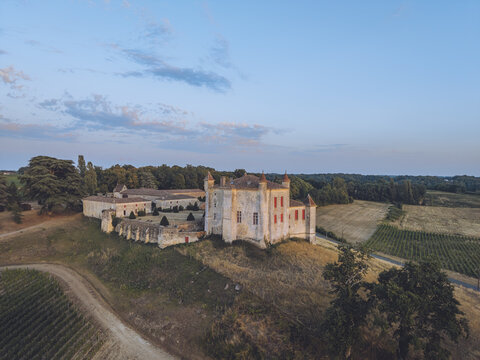 Aerial view of a historic chateau with stone walls and red shutters surrounded by vineyards and trees at sunset in Puisseguin, Nouvelle-Aquitaine, France.
