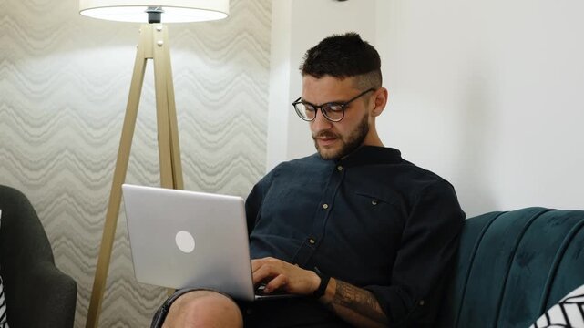 A young man wearing glasses works on his laptop while sitting comfortably on a sofa in a cozy modern living room. Digital worker in home office. Horizontal 4k footage