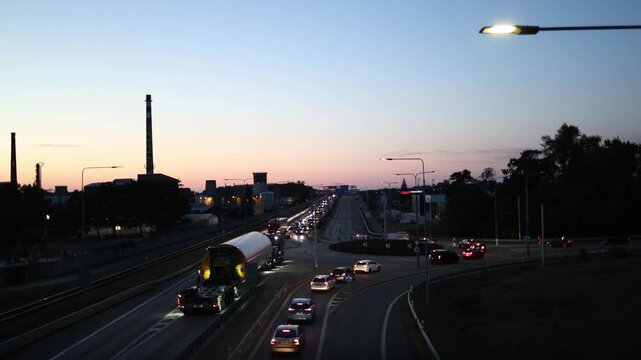 Oversized cargo transport at dusk while traffic is temporarily stopped as a heavy truck carrying a large industrial load slowly passes through a busy urban road junctions
