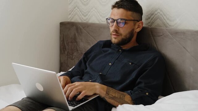Focused man wearing glasses works on laptop while lying in bed, remote work and modern lifestyle concept. Horizontal 4k footage