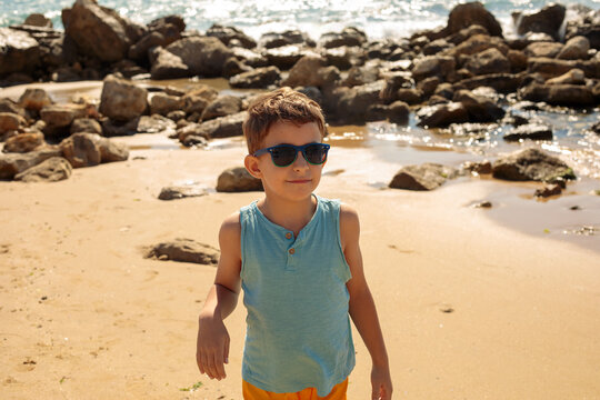 A young boy in sunglasses, a teal top, and orange shorts stands barefoot on a rocky beach shore, with dark boulders and gentle waves behind him