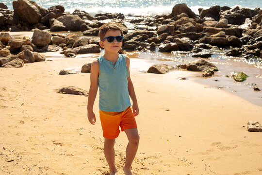 A young boy in sunglasses, a teal top, and orange shorts stands barefoot on a rocky beach shore, with dark boulders and gentle waves behind him