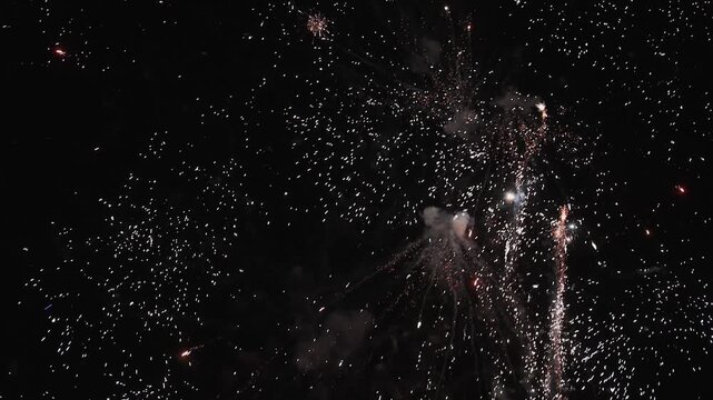 Spectacular fireworks explode across the night sky, sending bright colorful rockets and showers of glowing sparks upward through clouds of smoke. The dynamic display creates a festive atmosphere