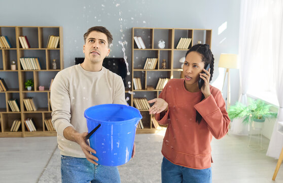 Couple handles water leak emergency with bucket. Man holds the bucket under a leaking ceiling as woman speaks via phone to a plumber for repair in apartment. Clear concept of home emergency.