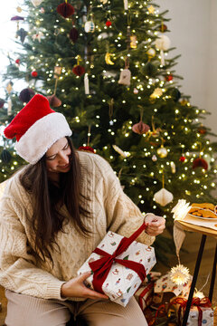 Happy woman wearing Santa hat opening red ribbon on Christmas gift box by glowing xmas tree in festive living room. Happy holidays