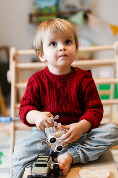 Cute toddler boy in red sweater sitting barefoot and playing with wooden sensory busy cube toy indoor in kid playroom