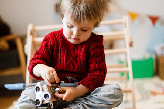 Focused toddler boy in red knitted sweater playing with wooden busy cube toy sitting indoor. Skill development