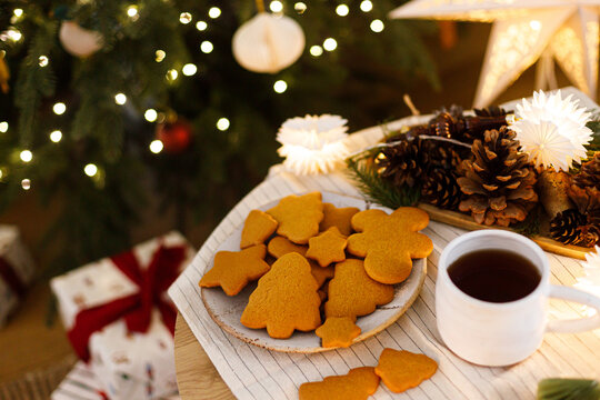 Top view of festive gingerbread cookies and warm cup of tea near illuminated Christmas tree. Festive background, cozy xmas eve