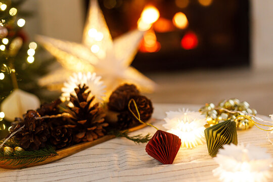 Festive christmas paper ornaments, pine cones, star garland lights on table and burning fireplace background. Cozy xmas eve. Winter holiday decorations