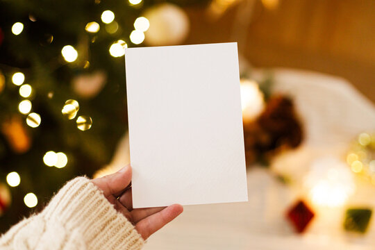 Hand Holding Blank Paper Card Mockup against Warm Glowing Christmas Tree Bokeh Lights Background. Festive Xmas card template with copy space