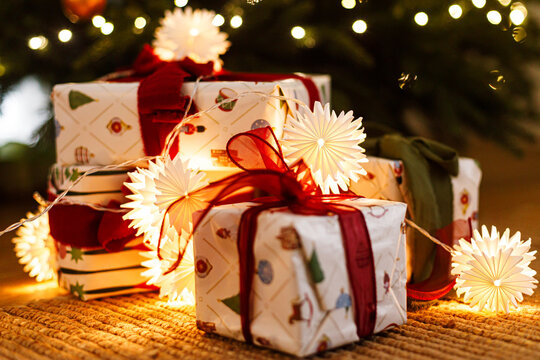 Close Up of Wrapped Christmas Gift Boxes Decorated with Red Ribbon and Illuminated Star Lights Under Xmas Tree. Merry Christmas