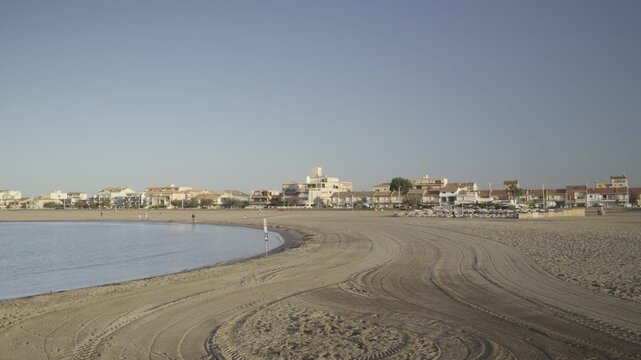 France, Valras Plage August 08, 2026 &mdash; Wide shot of a scenic sandy beach with tractor patterns on the sand and resort city buildings along the Mediterranean coast under a clear blue sky