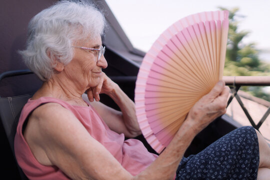 Elderly woman cooling herself with a fan while sitting on the balcony during a heatwave