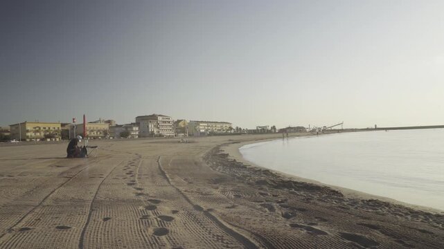 France, Valras Plage August 08, 2026 &mdash; Handheld shot of a wide sandy beach with a curved shoreline and calm sea bay, featuring a person sitting under an umbrella with resort buildings in the backgroun