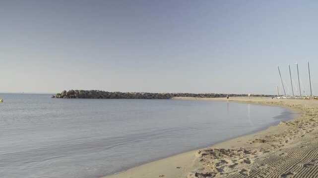 France, Valras Plage August 08, 2026 &mdash; Handheld shot of a peaceful sandy beach and calm sea bay with a rocky stone breakwater pier on the horizon and parked sports sailing catamarans
