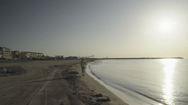 France, Valras Plage August 08, 2026 &mdash; Handheld shot of a athletic man jogging on a wide sandy beach along the calm sea shoreline during a bright vibrant golden hour sunrise with sun reflection on wat