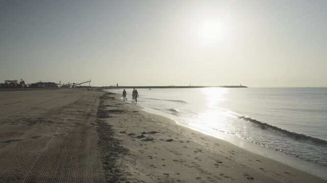 France, Valras Plage August 08, 2026 &mdash; Handheld shot of silhouettes of two people walking with a dog on a wide sandy beach shore during a bright golden sunrise with a strong sun reflection on water
